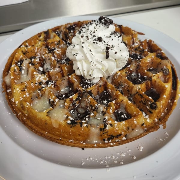 A golden waffle topped with whipped cream, chocolate cookie crumbles, and powdered sugar on a white plate.