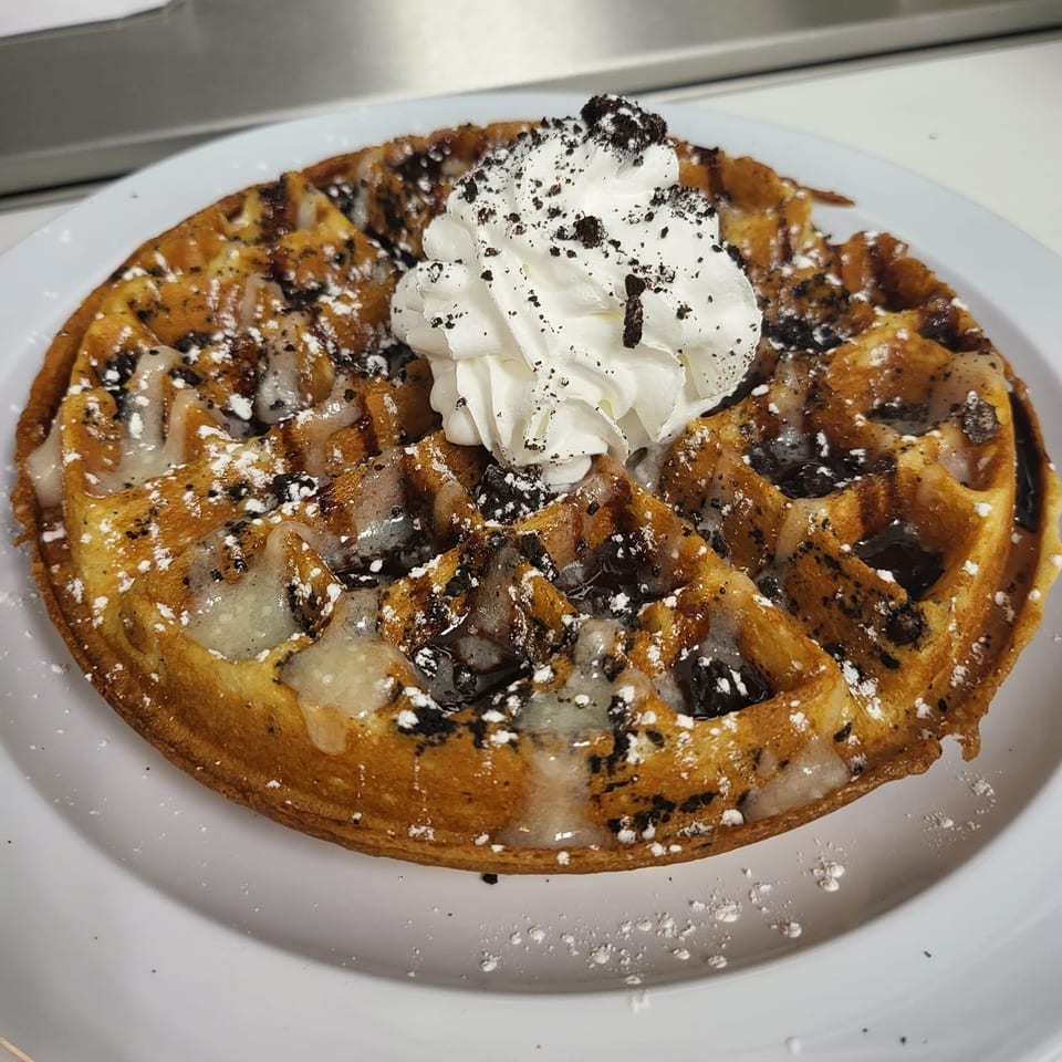 A golden waffle topped with whipped cream, chocolate cookie crumbles, and powdered sugar on a white plate.